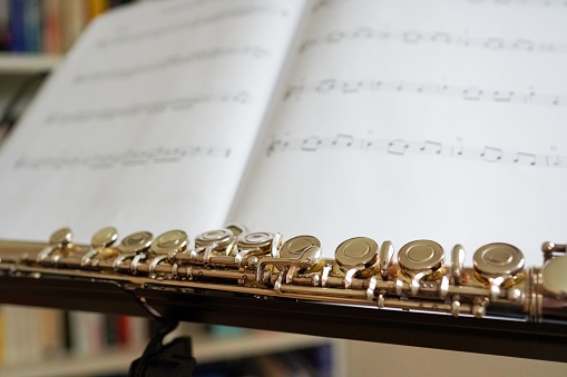 A closeup of a shiny flute against a musical score with a bookshelf in the background