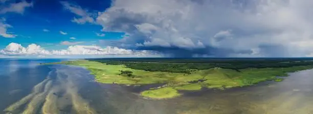 Aerial panoramic view to the shallow coastal sea with sandbanks, green grazed coastal meadow and inland marshes with passing rain front