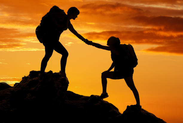 istock Silhouette, friends and helping while hiking during sunset on a mountain together against an orange sky background. Exercise in nature park climbing or trekking during a hike in nature during dusk 1443780971