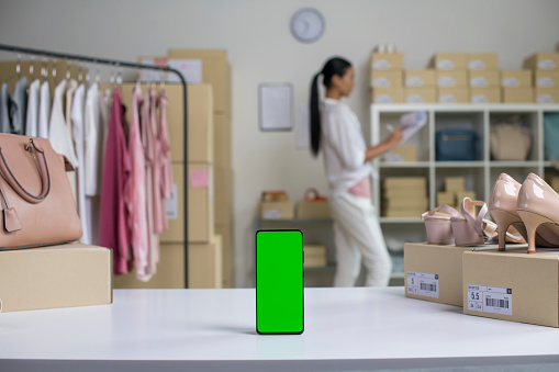 Smartphone with blank green screen standing on the desktop in the warehouses. Asian woman online seller checking stock and Inventory before sending to the customer.