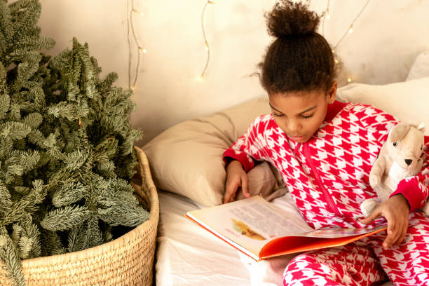 African-American girl in red and white pajamas is lying at home in bed and reading a book.Fir branches in the corner of the bedroom and garlands on the wall.New Year and Christmas concept