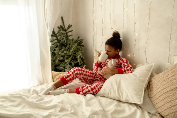 African-American girl in red and white pajamas is lying at home in bed and drinking cocoa.Fir branches in the corner of the bedroom and garlands on the wall.New Year and Christmas concept