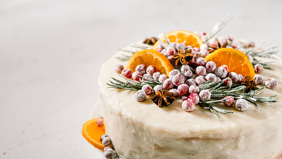 Winter homemade cake on white marble background. White cream christmas cake decored rosemary, cranberries, star anise and oranges slices. Copy space. Vertical