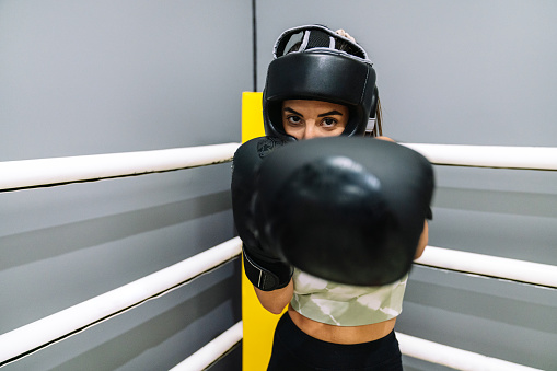 Front view of a young woman with boxing gloves and protective helmet throwing a punch to camera on a boxing ring.
