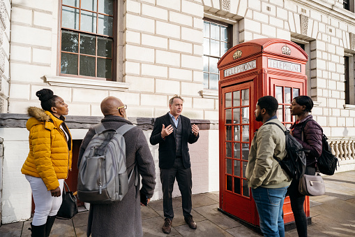 Mature Caucasian man and Black family in warm clothing standing next to iconic red telephone box as he explains history of Parliament Square.