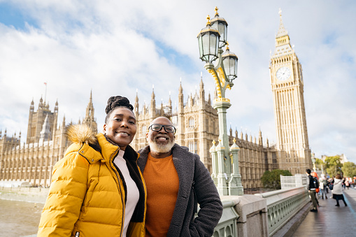 Waist-up view of tourists in warm clothing standing on Westminster Bridge, smiling at camera, with Big Ben and Houses of Parliament in background.