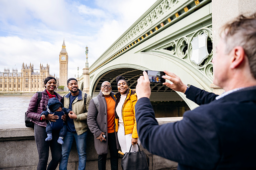 Front view of Black family with 4 month old baby smiling at smart phone held by tour guide with Westminster Bridge, River Thames, and landmarks in background.