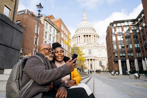 Vacationers in warm clothing sitting outdoors on steps and smiling at smart phone as they capture travel memory, St. Paul’s Cathedral in background.
