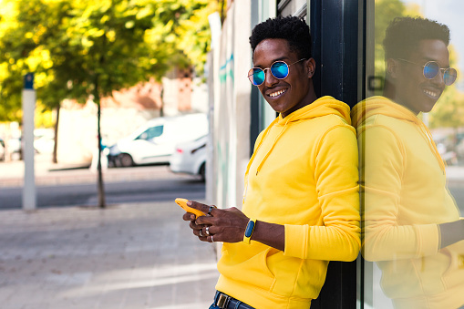 A smiling young African man is leaning against the wall of a building in the street next to the glass of a shop window in which he is reflected. The guy is holding his cell phone.
