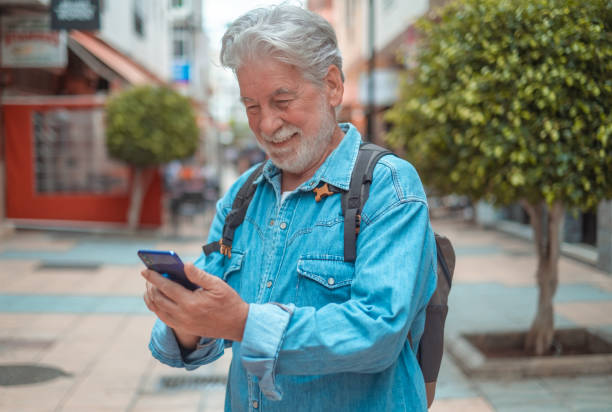 istock Handsome senior man walking in the city wearing backpack and using phone. White haired caucasian grandparent enjoying free time and technology 1443822514