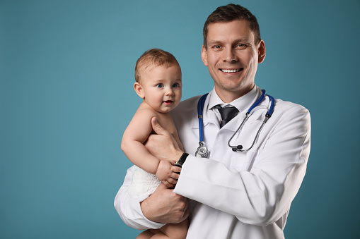 Pediatrician with cute little baby on light blue background