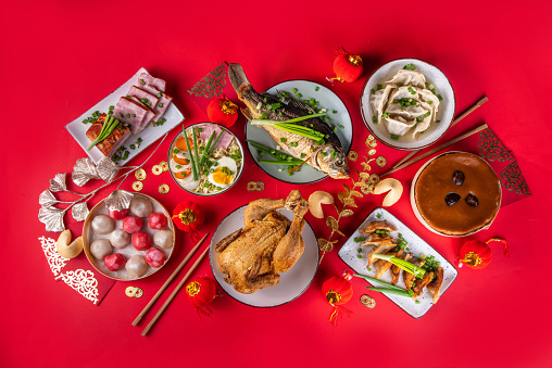 Traditional Chinese lunar New Year dinner table, party invitation, menu background with pork, fried fish, chicken, rice balls, dumplings, fortune cookie, nian gao cake, noodles, chinese decorations