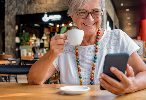 Smiling senior woman relaxing for a break at coffee shop with chocolate cake and coffee cup. Carefree elderly woman with eyeglasses and necklace using mobile phone