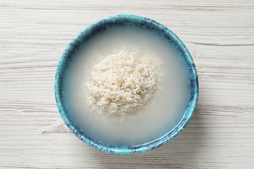 Bowl with rice soaked in water on white wooden table, top view