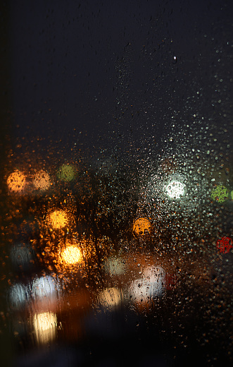 Bright lights of night city through windows glass in drops of rain, abstract background with blurred  lights and bokeh in rainy weather