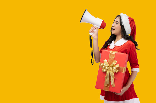 Woman wearing red Santa Claus outfit and Santa Claus Hat shouting loud to copy space holding a megaphone gift box Yellow background Christmas x-mas winter happiness holiday and party concept