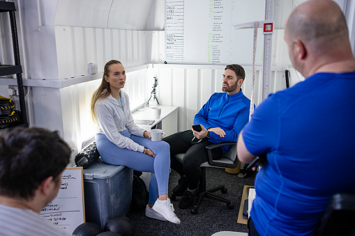 An over-the-shoulder, medium shot of a group of colleagues wearing sports clothing in their gym office on a winter's day. They are gathered around having a meeting.