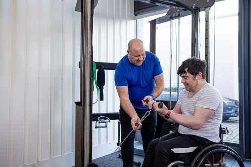 A wide shot of a personal trainer and his client wearing sports clothing in a gym on a winter's day. His client uses a wheelchair and has a Continuous Glucose Monitor on his arm. The client is using a cable machine to do bicep curls while his trainer gives him guidance and motivation.