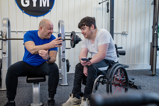 A medium shot of a personal trainer and his client wearing sports clothing in a gym on a winter's day. His client is using a wheelchair and has a Continuous Glucose Monitor on his arm. They are taking a break from working out and the trainer is showing his client something on his smartphone.