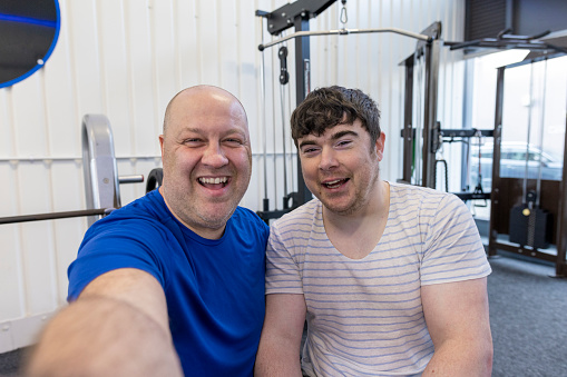 A selfie shot of a personal trainer and his client wearing sports clothing in a gym on a winter's day. His client is using a wheelchair and has a Continuous Glucose Monitor on his arm. They are taking a selfie on a smartphone.