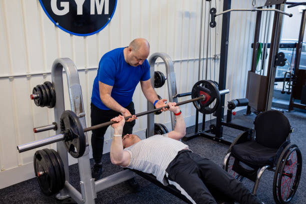 Helping My Client A wide shot of a personal trainer and his client wearing sports clothing in a gym on a winter's day. His client uses a wheelchair and has a Continuous Glucose Monitor on his arm. The client is doing chest presses on a weight bench while his trainer gives him guidance and motivation. bench press spotter stock pictures, royalty-free photos & images