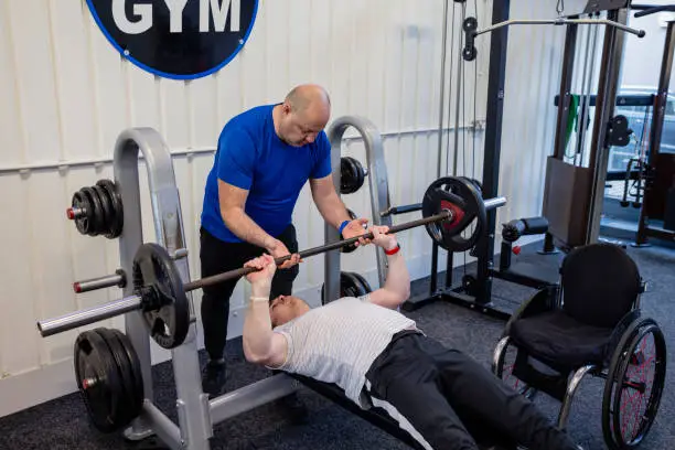 A wide shot of a personal trainer and his client wearing sports clothing in a gym on a winter's day. His client uses a wheelchair and has a Continuous Glucose Monitor on his arm. The client is doing chest presses on a weight bench while his trainer gives him guidance and motivation.