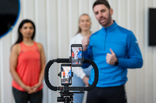 A medium shot of a personal trainer and two of his clients wearing sports clothing in a gym on a winter's day. He is live streaming a fitness class with two smartphones set up on a tripod.