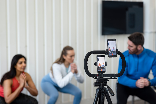 A medium shot of a personal trainer and two of his clients wearing sports clothing in a gym on a winter's day. He is live streaming a fitness class with two smartphones set up on a tripod.