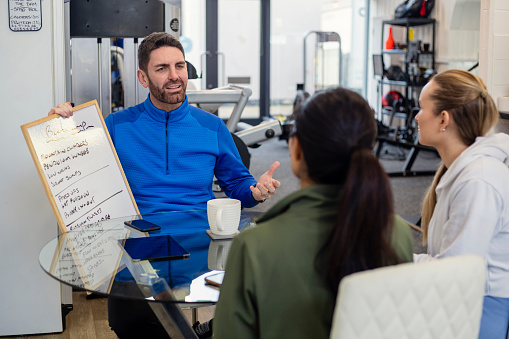 An over-the-shoulder, medium shot of a personal trainer and two of his clients wearing sports clothing in a gym on a winter's day. They are sitting around a table, talking and having a cup of coffee. The personal trainer is showing his clients a whiteboard with a BootCamp training plan on.