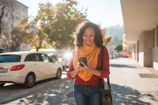 Dark-haired, pretty Latina woman with yellow scarf, walking and texting on a city street while smiling. 3/4 length image. Looking away. Cars in the background.