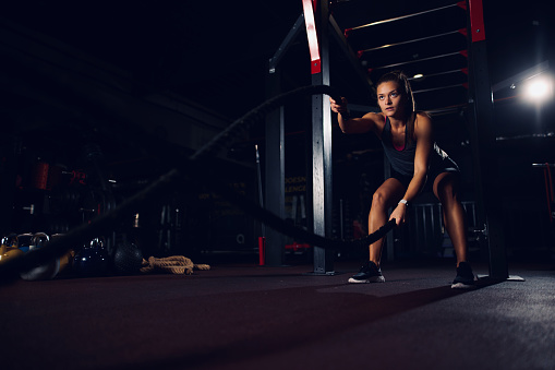 Young fit sportswoman working out with battle ropes at gym