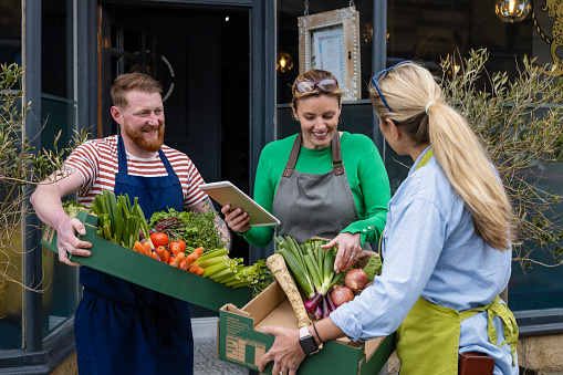 A front-view shot of a man and woman holding a bunch of fresh vegetables which includes tomatoes, beetroots, carrots radishes, cabbage, horseradish and celery. They are buying crates of vegetables for the restaurant.