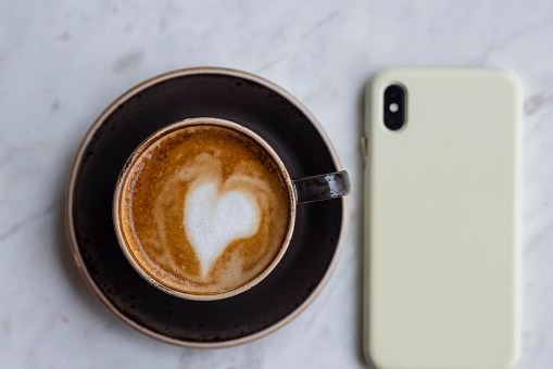 A directly above shot of a freshly made cup of coffee with heart shaped froth art on a table with a smartphone.