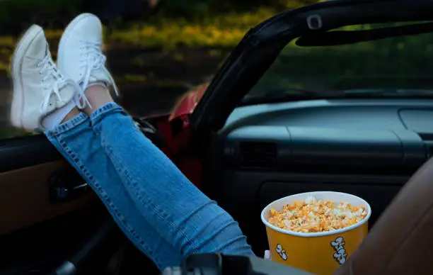 Woman hanging her legs out of the red cabriolet car while watching a movie with popcorn at drive in cinema. Close up image with selective focus. Entertainment, leisure activities, hobby concept.