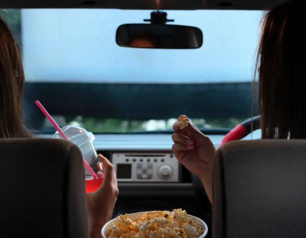 Two female friends with popcorn sitting in the car while watching a movie at drive in cinema. Selective focus. Entertainment, leisure activities, hobby concept.