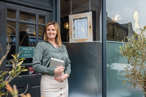 A front-view three-quarter-length portrait of a mid-adult woman, smiling and looking at the camera while holding a digital tablet, she is standing in front of her new small business restaurant in Northumberland, England.