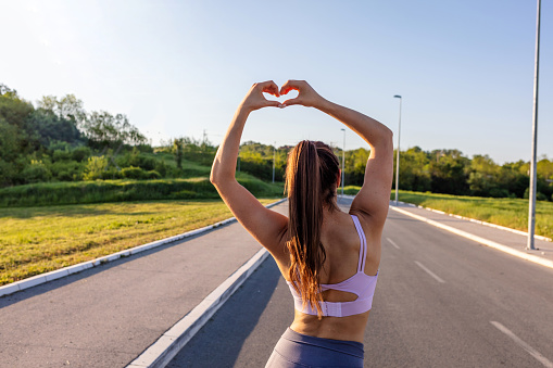 Rear view portrait of a sporty young woman making a heart shape with her hands while exercising outdoors.