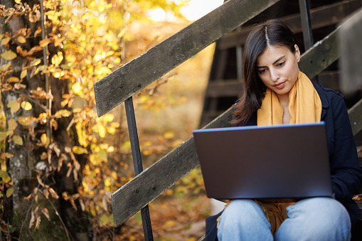 Beautiful young woman sitting on wooden staircase and using laptop in autumn nature