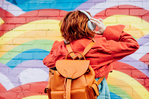 Back view stylish woman in bright clothes with backpack wearing wireless headphones and listening to music while looking on rainbow graffity on the brick wall. Fashionable hipster lifestyle
