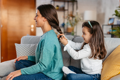 Young daughter braiding her mother's hair on the sofa in the living room.