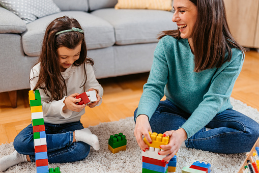 Young mother and her daughter playing with brick toys on the carpet in the living room.