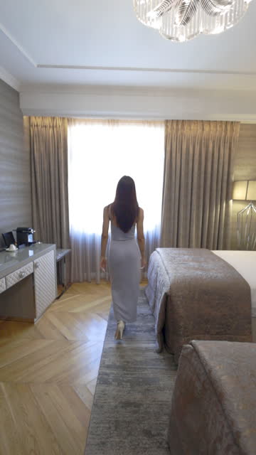 A woman in a dress watches the view from the hotel window, Young woman opening the curtain in the hotel room and watching the view