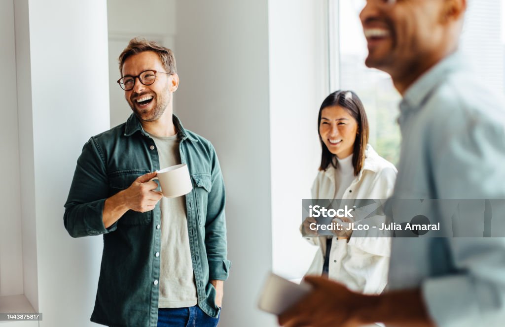 Happy business people taking a coffee break at work Business people laughing together during a coffee break at work. Group of young professionals standing together in an office. Coffee - Drink Stock Photo