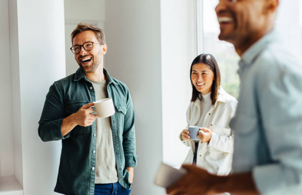 Happy business people taking a coffee break at work Business people laughing together during a coffee break at work. Group of young professionals standing together in an office. casual clothing stock pictures, royalty-free photos & images