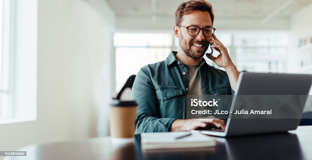 Software designer speaking to his client on the phone in an office Software designer discussing a new project with his client on the phone. Creative business man working on a laptop in an open plan office. Using Phone Stock Photo