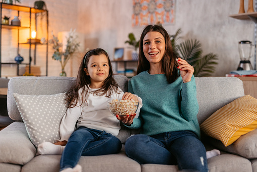 Young mother and her daughter sitting on the sofa in the living room, watching a funny movie and eating popcorn.