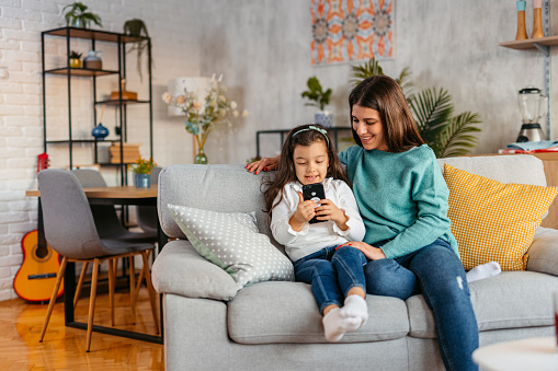 Young mother and her daughter using smart phone together on the sofa in the living room.