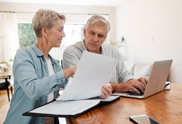 istock Senior couple, laptop and home finance while reading paperwork documents and typing on computer for retirement savings, mortgage and online banking. Mature man and woman doing bill payment or booking 1443879043