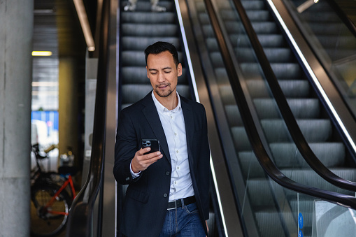 Confident Asian Businessman Using The Escalator While Working On His Smartphone