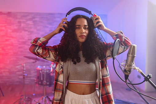 A portrait of an attractive curly haired black female singer putting on her headphones before recording a song in a beautiful music studio. She is looking at the camera.There is a microphone besides the beautiful singer and pink and purple lights behind her. The drums in the back are partially visible. The black musician looks confident and serious. She has beautiful long curly hair with a lot of volume.